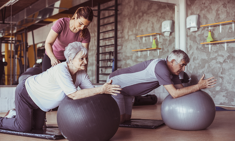 A woman with gray hair and a man with gray hair are shown each leaning into a yoga ball with their forearms as they kneel on the ground, and the woman is being assisted by a standing woman. This illustrates “prehab,” or rehabilitation exercises done before surgery to improve outcomes.
